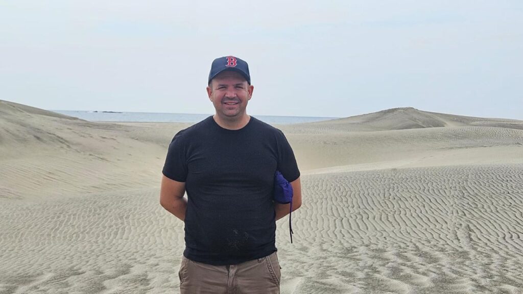 Héctor Muskus, encargado de Operaciones en Guardia Consultores, sonriendo en un paisaje de dunas de arena junto al mar en Santo Domingo Ingenio, Oaxaca.