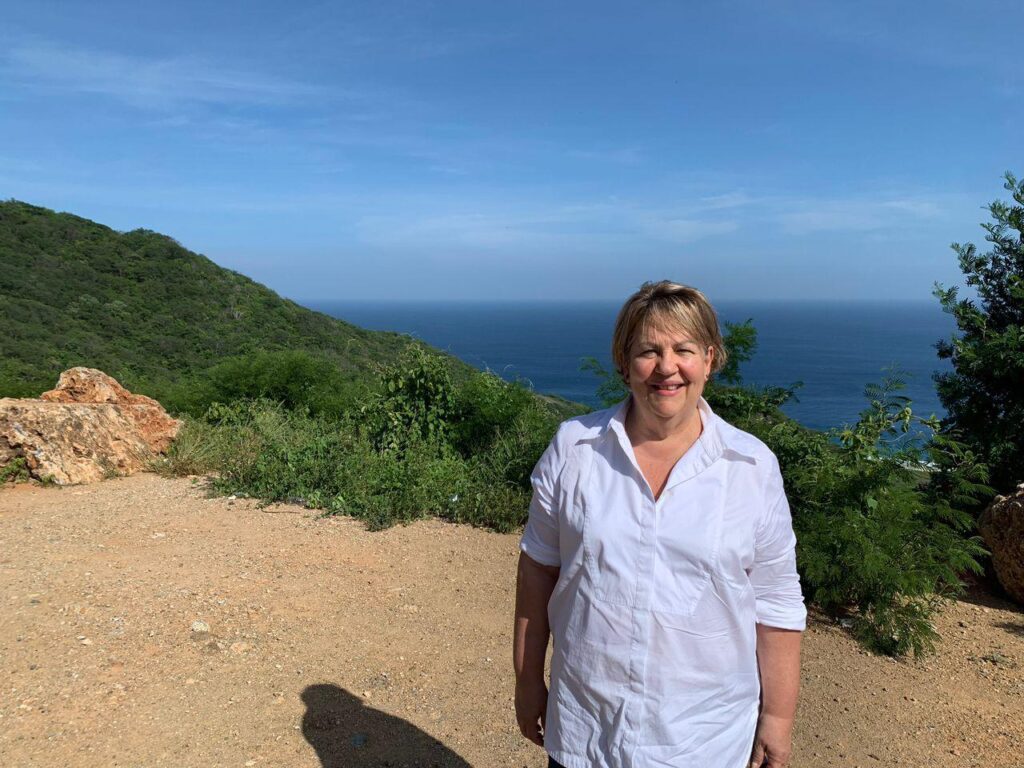 Inés Guardia, directora de Guardia Consultores, sonriendo al aire libre con un fondo de colinas verdes y el océano en Santo Domingo Ingenio, Oaxaca.