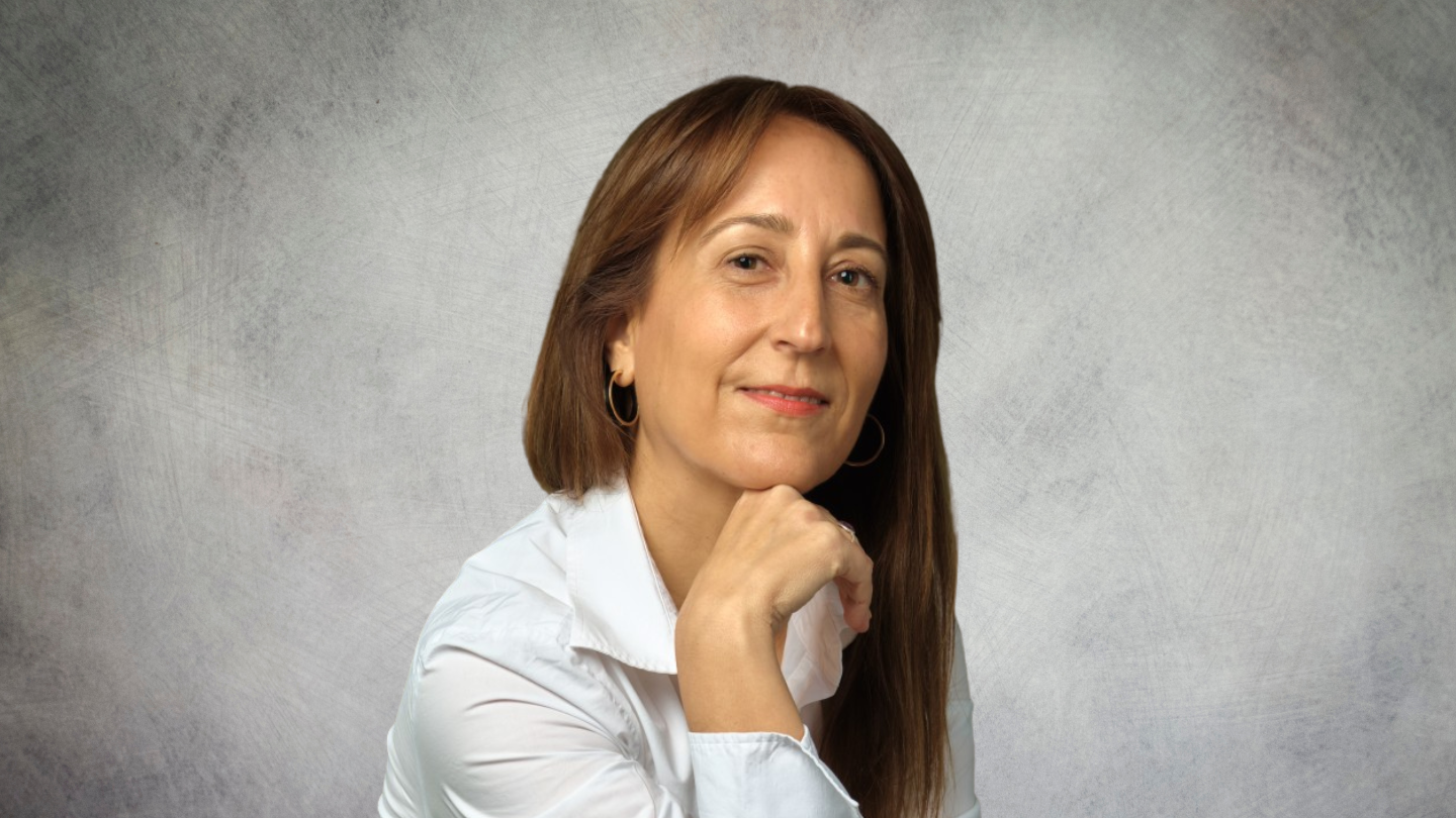 Retrato de Belén Merino, una mujer de cabello castaño claro, vestida con una camisa blanca, mirando a la cámara con una expresión serena y profesional. Su mano descansa bajo su barbilla, en una pose de reflexión. Fondo neutro.