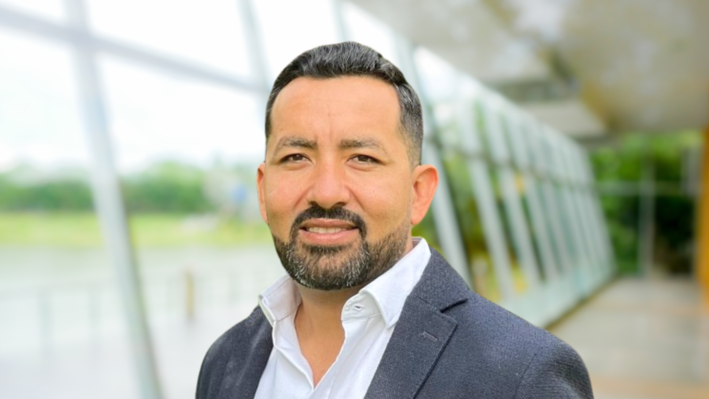 Retrato profesional de David Parra, vistiendo un saco formal y camisa blanca, con un fondo arquitectónico moderno y vegetación desenfocados.